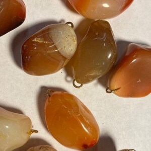 May include: A collection of eight carnelian gemstone pendants with gold-toned metal loops. The carnelian stones are various shades of orange and brown, with some having white bands.