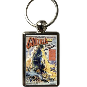 May include: A silver metal keychain featuring a vintage Godzilla movie poster. The poster is red, yellow, and blue with the text "Raging Through the World on a Rampage of Destruction! Godzilla It's Alive! King of the Monsters! Awesome! Raymond Burr"