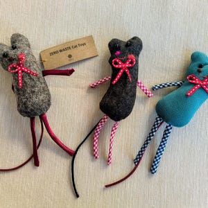Puede incluir: Tres juguetes para gatos hechos a mano de fieltro con forma de ratón. Los ratones son de diferentes colores: gris, negro y azul verdoso. Cada ratón tiene un lazo rojo y patas a cuadros blancos y rojos. Los juguetes están etiquetados como "ZERO WASTE Cat Toys".