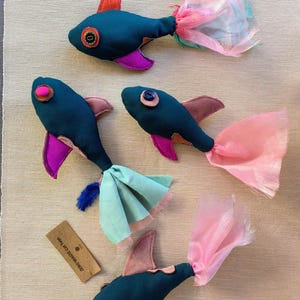 Puede incluir: Cinco juguetes de pescado de tela azul turquesa con detalles en rosa, rojo y naranja. Cada pez tiene una cola de tela diferente, incluyendo tul rosa, tul azul y una combinación de tul rosa y azul. Los peces tienen ojos de botón y el texto "ZERO WASTE CRAFT" está en una etiqueta marrón.