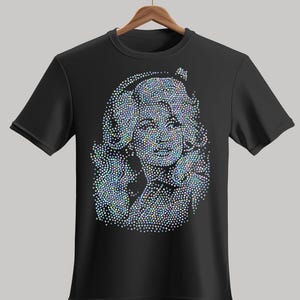 May include: A black short-sleeved t-shirt featuring a graphic design composed of iridescent, multi-coloured rhinestones that form the portrait of a person with voluminous hair. The text GlittArt.com is printed in grey at the bottom of the image.