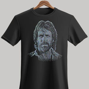 Könnte beinhalten: Schwarzes T-Shirt mit Rundhalsausschnitt und einem Porträt eines Mannes, das aus schillernden Punkten besteht. Das Design ist auf dem T-Shirt zentriert, das an einem Holzbügel hängt.