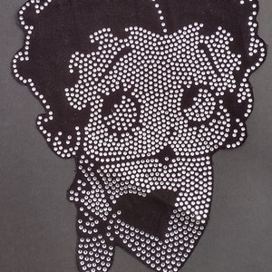Puede incluir: Un parche de tela negro con un diseño de rostro de mujer en strass. Los strass son transparentes y crean un efecto brillante.