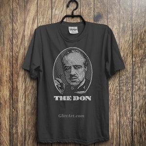 Può includere: Una maglietta grigio scuro con un design in strass bianco di un viso maschile in un cerchio. Il testo "The Don" è sotto l'immagine e il sito web "GlittArt.com" è sotto il testo.