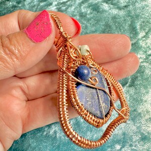 May include: A copper wire wrapped pendant featuring a blue lapis lazuli stone with gold flecks. The pendant is accented with a small white bead and a copper wire loop.