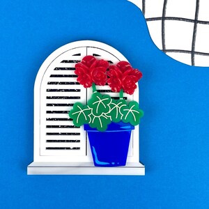 May include: A white window with a blue pot of red flowers and green leaves. The window has white shutters and is set against a blue background.