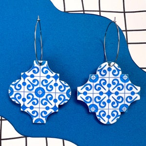 Puede incluir: Un par de pendientes de aro con estampado azul y blanco. Los pendientes están hechos de acrílico blanco con un patrón geométrico azul. Los aros son plateados.