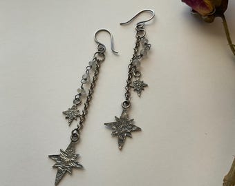 May include: A pair of dangling earrings featuring dark oxidized metal chains, small translucent beads, and textured star-shaped charms. Each earring hangs from a curved ear wire, showcasing a layered design with two distinct star sizes.