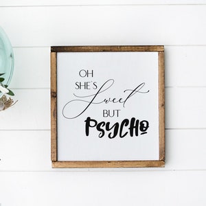 May include: A white wooden sign with a brown frame, featuring the text "Oh she's sweet but psycho" in black lettering.