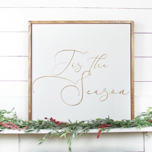 May include: A white wooden sign with a gold inscription that reads "Tis the Season" hanging above a white fireplace mantel decorated with greenery.