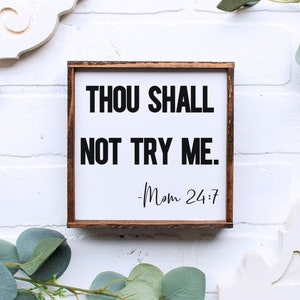K&ouml;nnte beinhalten: Ein wei&szlig;es Holzschild mit braunem Rahmen, auf dem der Text "Thou Shall Not Try Me. -Mom 24:7" in schwarzen Buchstaben steht.