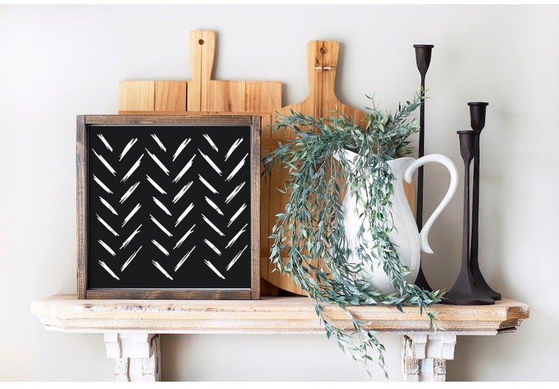May include: A black and white framed sign with a chevron pattern. The sign is on a shelf with a white pitcher, greenery, and candlesticks.