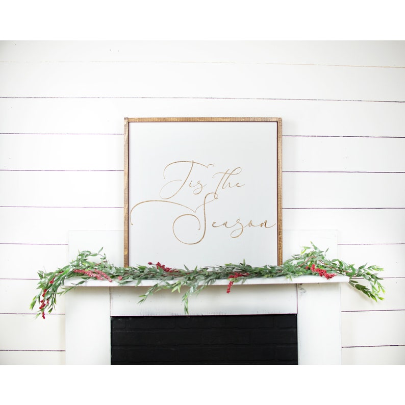 May include: A white wooden sign with a gold inscription that reads "Tis the Season" hanging above a white fireplace mantel decorated with greenery.