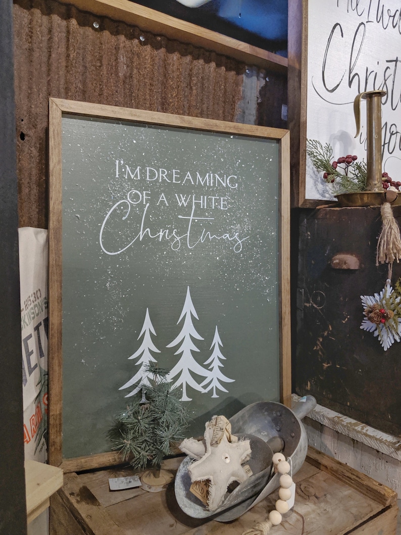 K&ouml;nnte beinhalten: Ein Holzschild mit gr&uuml;nem Hintergrund und wei&szlig;en Buchstaben, auf dem "Ich tr&auml;ume von einem wei&szlig;en Weihnachten" steht. Das Schild hat einen Schneeeffekt und drei wei&szlig;e Silhouetten von B&auml;umen.