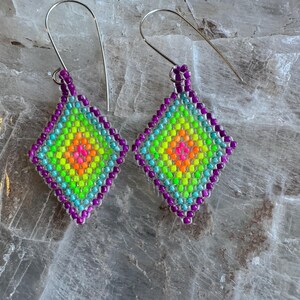 Peut inclure: Une paire de boucles d'oreilles en forme de losange. Les boucles d'oreilles présentent un dégradé de couleurs vives, allant du rose vif au centre à l'orange, au vert et au turquoise, souligné de perles violettes. Les crochets d'oreilles sont argentés.