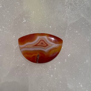 Op de afbeelding: Een halfronde agaat cabochon met een rood, oranje en wit bandpatroon. De agaat heeft een glad, gepolijst oppervlak.