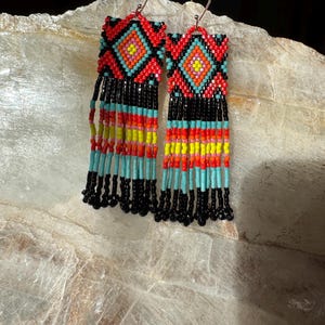 Puede incluir: Pendientes de cuentas hechos a mano con un diseño geométrico. Los pendientes presentan un patrón rojo, naranja y negro en la parte superior, con largas franjas en negro, turquesa, amarillo y rojo. Cuelgan de ganchos plateados.
