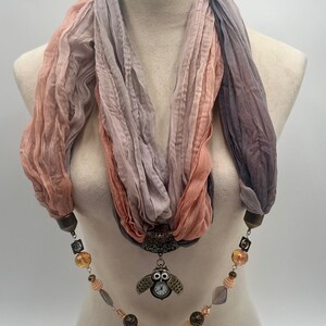 May include: A peach, lavender, and grey gradient scarf displayed on a mannequin. The scarf has a pendant with an owl design and a beaded necklace with various shapes and colours.