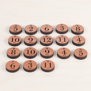 Puede incluir: Piezas de juego de madera con letras de la A a la R y n&uacute;meros del 2 al 12. Las piezas est&aacute;n dispuestas en semic&iacute;rculo sobre una superficie de madera clara.