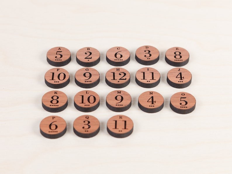 Puede incluir: Piezas de juego de madera con letras de la A a la R y n&uacute;meros del 2 al 12. Las piezas est&aacute;n dispuestas en semic&iacute;rculo sobre una superficie de madera clara.