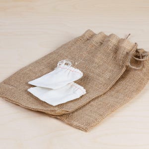 Puede incluir: Dos peque&ntilde;as bolsas de tela blanca est&aacute;n anidadas dentro de un gran saco de arpillera. El saco est&aacute; hecho de arpillera marr&oacute;n natural y tiene un cierre de cord&oacute;n.