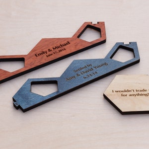 Puede incluir: Dos llaveros de madera con forma de casa con texto grabado. El llavero superior es rojo y dice "Emily & Michael June 17, 2018". El llavero inferior es azul y dice "Settled by Amy & David Young 6.24.14". Tambi&eacute;n se muestra un llavero hexagonal de madera con el texto "&iexcl;No te cambiar&iacute;a por nada!"