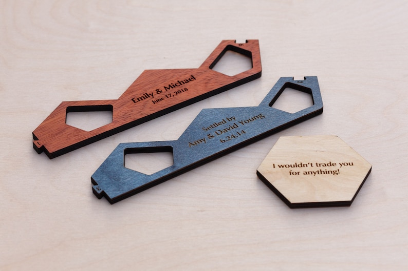 Puede incluir: Dos llaveros de madera con forma de casa con texto grabado. El llavero superior es rojo y dice "Emily & Michael June 17, 2018". El llavero inferior es azul y dice "Settled by Amy & David Young 6.24.14". Tambi&eacute;n se muestra un llavero hexagonal de madera con el texto "&iexcl;No te cambiar&iacute;a por nada!"