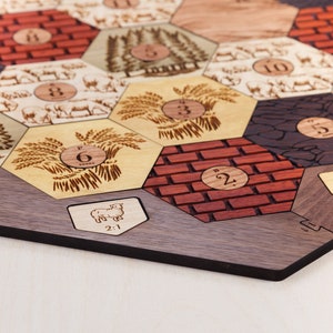 Puede incluir: Un tablero de juego de madera con fichas hexagonales que presentan diferentes im&aacute;genes y n&uacute;meros. Las fichas incluyen im&aacute;genes de trigo, ovejas, madera, ladrillos y piedra. Los n&uacute;meros en las fichas van del 1 al 6. El tablero est&aacute; hecho de madera oscura y tiene un borde negro.