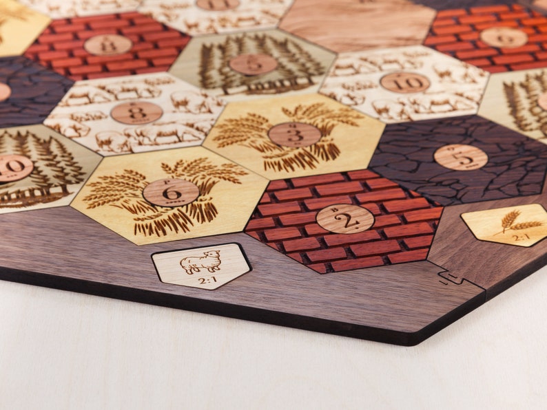 Puede incluir: Un tablero de juego de madera con fichas hexagonales que presentan diferentes im&aacute;genes y n&uacute;meros. Las fichas incluyen im&aacute;genes de trigo, ovejas, madera, ladrillos y piedra. Los n&uacute;meros en las fichas van del 1 al 6. El tablero est&aacute; hecho de madera oscura y tiene un borde negro.