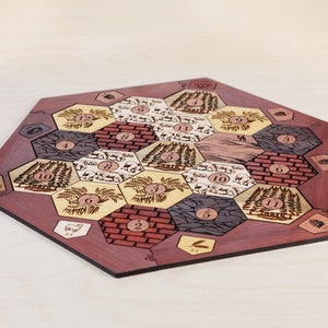 Puede incluir: Un tablero de juego hexagonal de madera con un borde de caoba roja. El tablero est&aacute; dividido en 19 fichas hexagonales, cada una con un tipo de recurso diferente: trigo, ovejas, madera, ladrillo y mineral. Cada ficha tiene un n&uacute;mero del 2 al 12.