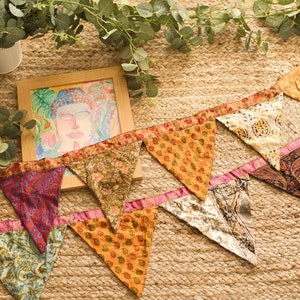 May include: A colourful fabric bunting with triangular flags featuring various patterns and colours, including pink, orange, blue, and green. The bunting is strung on a jute background.