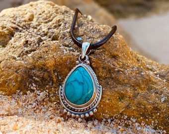 Puede incluir: Un collar con un colgante de piedra turquesa en forma de lágrima con engaste de plata. El colgante está colgado de un cordón negro. El collar está apoyado en una roca con arena en el fondo.