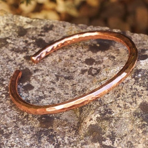 Peut inclure: Un bracelet en cuivre martelé avec une finition lisse et polie. Le bracelet est posé sur une roche rugueuse et texturée.