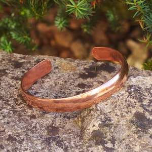 Puede incluir: Un brazalete de cobre simple con una superficie lisa y redondeada. El brazalete está sobre una roca gris y rugosa.