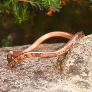 Pode incluir: Um bracelete de cobre com um design ondulado e gravuras florais. O bracelete está apoiado numa rocha castanha.