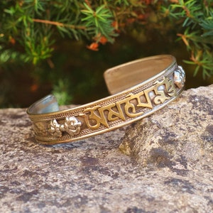 Puede incluir: Un brazalete de plata y oro con un mantra tibetano inscrito en él. El brazalete está sobre una roca gris.