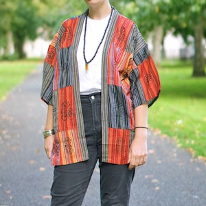 May include: A colourful patchwork kimono jacket in orange, red, black, and grey. The jacket is worn over a white t-shirt and dark grey jeans. The outfit is completed with high-top trainers with a black and white pattern.