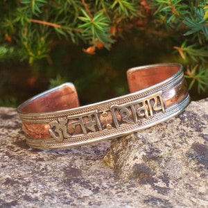 Puede incluir: Una pulsera de puño de cobre y plata con texto sánscrito grabado. La pulsera está abierta en los extremos y descansa sobre una piedra gris, con follaje verde en el fondo. El diseño de la pulsera presenta una combinación de metal pulido y texturizado.