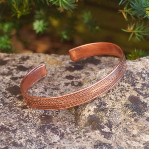 Op de afbeelding: Een koperen armband met een gevlochten ontwerp. De armband ligt op een grijze steen.