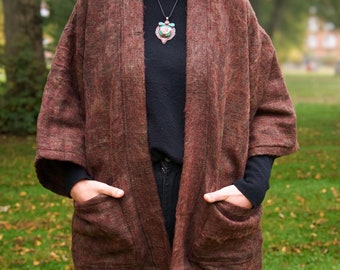 Puede incluir: Un kimono de lana marrón con mangas largas y dos bolsillos. El kimono se lleva sobre un jersey negro y unos vaqueros. La persona lleva un collar con un colgante de piedra turquesa.