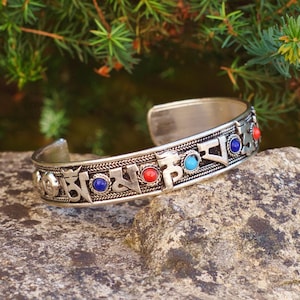 May include: Silver cuff bracelet with engraved symbols and colorful gemstone accents. The bracelet features blue, red, and turquoise stones. The bracelet is displayed on a gray stone with a green leafy background.