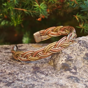 Op de afbeelding: Een gevlochten koperen en zilveren armband met een geweven ontwerp. De armband ligt op een ruwe, grijze rots.