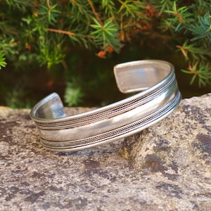 May include: A silver cuff bracelet with a textured design. The bracelet is resting on a grey stone, with green foliage in the background. The cuff has a simple, elegant design.