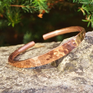May include: A copper cuff bracelet with a hammered texture. The open-ended bracelet is a warm, reddish-brown color and sits on a gray stone. The background features green foliage.