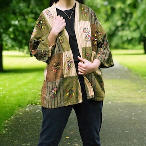 May include: A patchwork kimono jacket in olive green, brown, and beige, with embroidered floral patterns. The jacket is worn over a black top and paired with dark blue jeans. The background shows a green park setting.