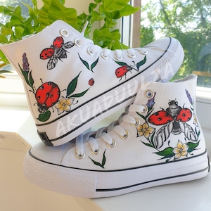 Könnte beinhalten: Weiße High-Top-Canvas-Schuhe mit handbemalten Marienkäfern, Blumen und Blättern. Die Schuhe haben schwarze Schnürsenkel und einen schwarzen Besatz um die Sohle.