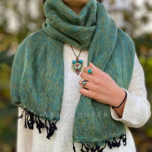 May include: A green and blue pashmina shawl with black fringe. The shawl is draped around a person's neck and shoulders. The person is wearing a white sweater and has a turquoise pendant necklace on.