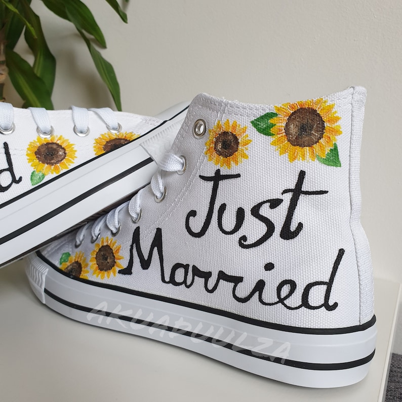 May include: White high-top canvas shoes with black laces and a black trim around the sole. The shoes are painted with sunflowers and the words "Just Married".