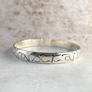 Puede incluir: Pulsera de plata con un diseño grabado. El diseño es una serie de símbolos y puntos.