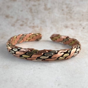 Puede incluir: Un brazalete de cobre trenzado con un diseño tejido en tonos de cobre, latón y plata.
